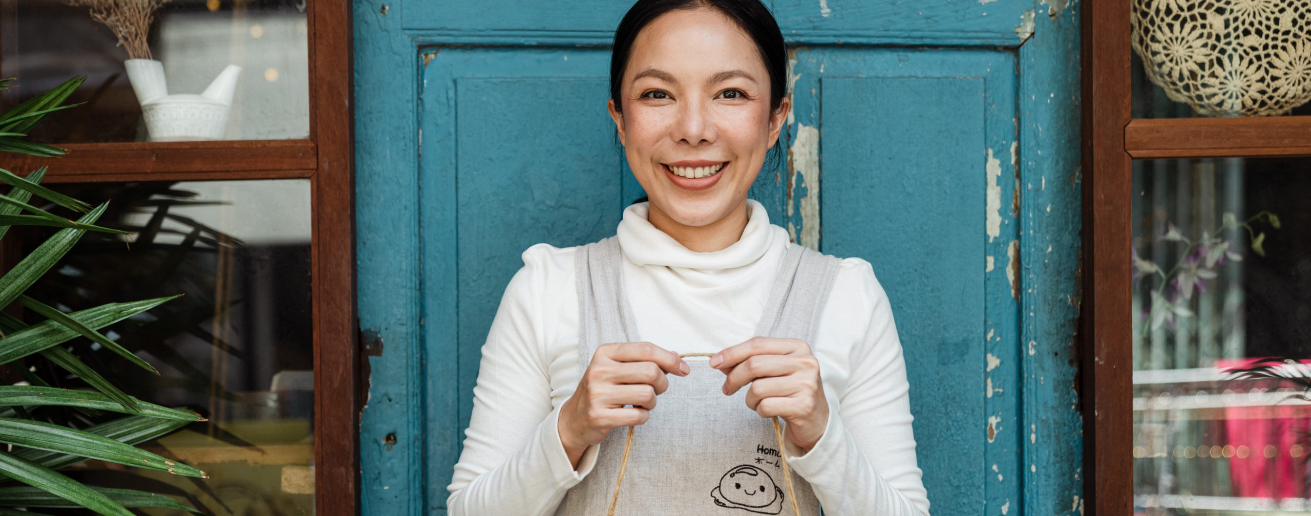 A woman with dark hair, a white turtleneck, and a gray apron smiles while standing in front of a turquoise door. 						