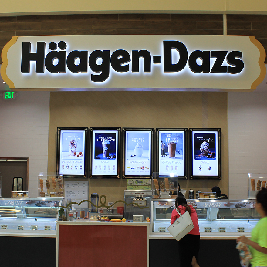 A full view of a Haagen-Dazs storefront including customers, dipping cabinets, and video menu displays hung on a back wall.