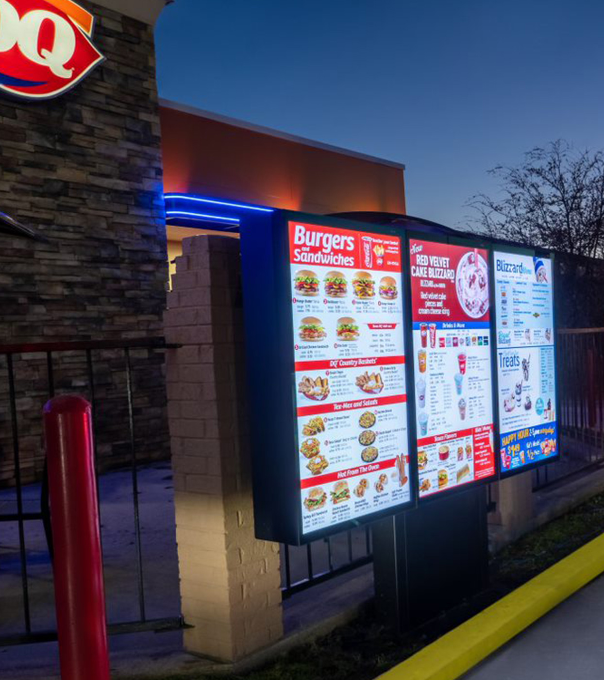 A brightly lit Dairy Queen drive-thru menu board against the restaurant's brick exterior. 							