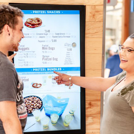 Two people are smiling and pointing toward menu items on a digital menu board for Auntie Anne's pretzels. 							