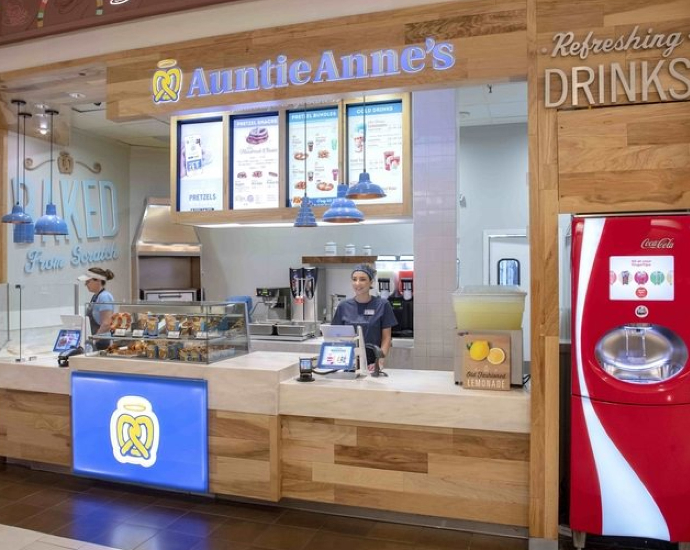 A smiling cashier stands at the counter at an Auntie Anne's pretzels witha vibrant menu in the background.							