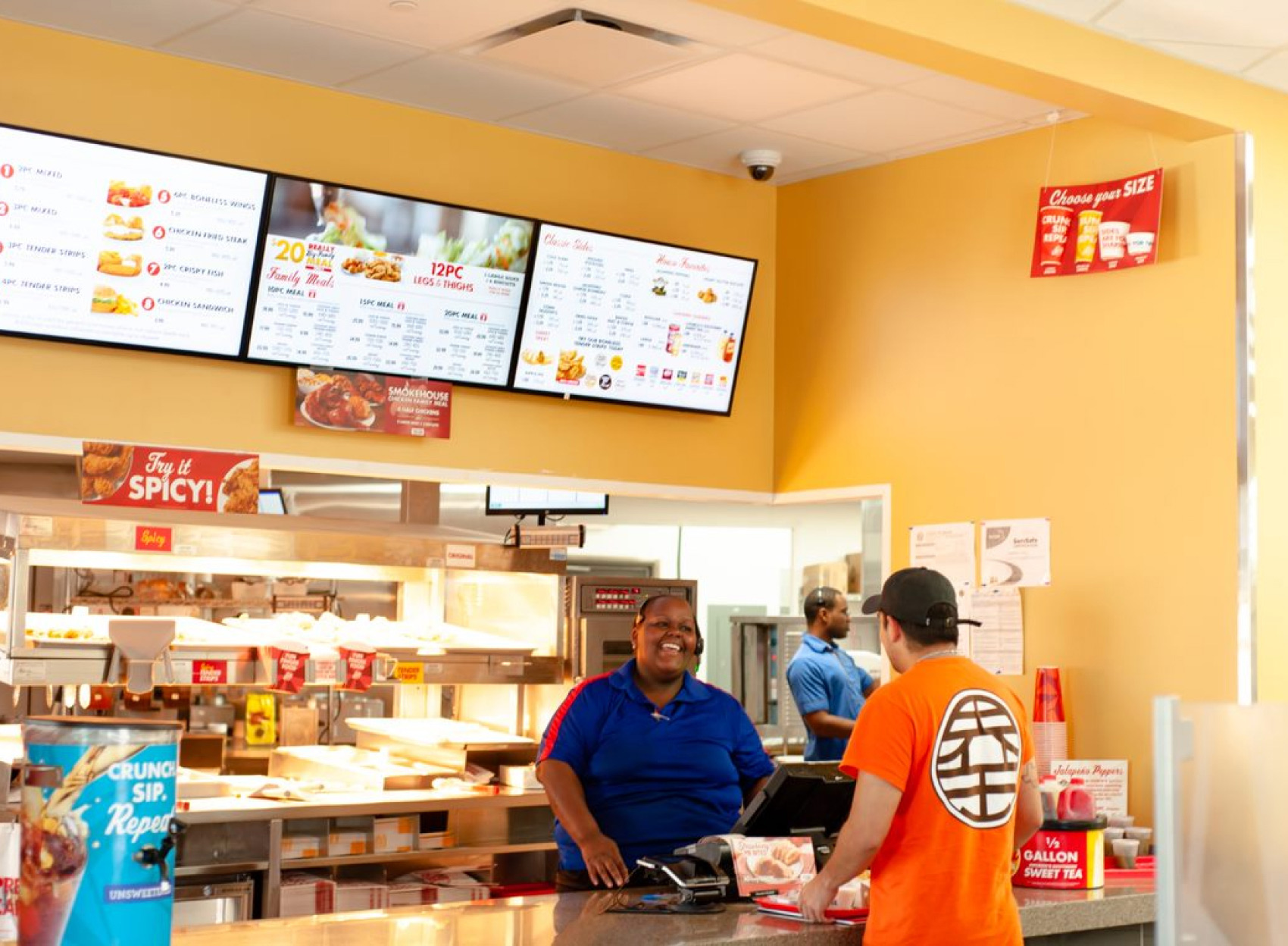 A smiling cashier helps a customer at the counter with branded signage and three digital menu displays behind them. 							